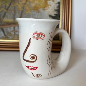 May include: White ceramic mug with a face design. The mug features a brown nose, red lips, and an eye with red and brown details. The handle curves from the top of the mug. The mug is on a white surface.