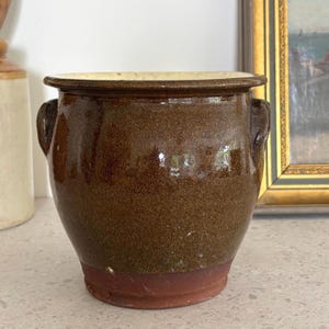 May include: A brown glazed ceramic pot with two small handles. The pot has a wide rim and a reddish-brown base. The interior rim is a light cream colour. The pot is likely used for planting or storage.
