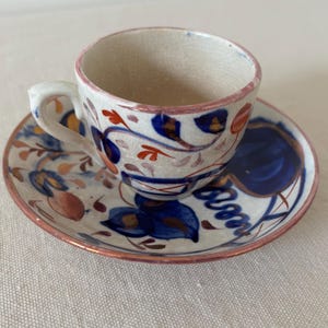 May include: A vintage teacup and saucer set with a floral pattern in shades of blue, red, and gold. The cup and saucer have a white background with a pink rim.