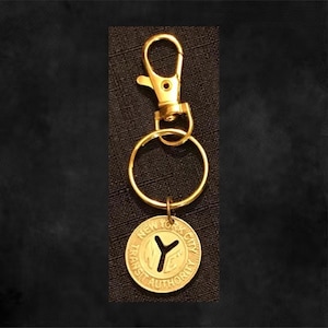 May include: A gold-colored keychain featuring a New York City Transit Authority token. The token has a cut-out "Y" shape and is attached to a gold ring and clasp. The keychain is designed for keys or accessories.