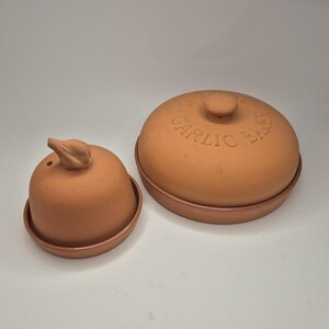 May include: Two terracotta garlic bakers. One is a small, round baker with a lid shaped like a garlic clove. The other is a larger, round baker with a lid and the words "ORIGINAL GARLIC BAKER" embossed on the top.