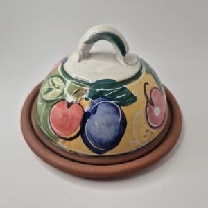 May include: A ceramic cheese dish with a domed lid and a terracotta-coloured base. The lid is decorated with colourful fruit illustrations, including red apples and blue plums, set against a yellow background. The handle is white with green accents.