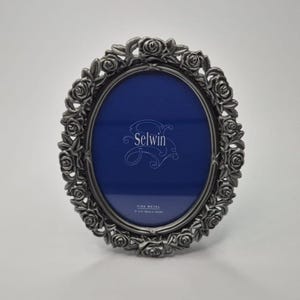 May include: An oval-shaped picture frame made of dark grey metal with a decorative border of sculpted roses. The centre contains a blue insert with the text Selwin, Fine Metal, and 5 x 7 inches (13cm x 18cm) printed in white lettering.