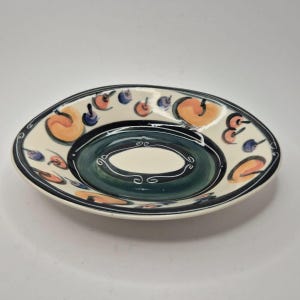 May include: Oval ceramic serving dish with a cream interior and a dark green centre. The rim is decorated with orange, blue, and black painted designs. The dish has a black border.
