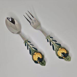 May include: A stainless steel spoon and fork set. The handles feature a yellow fruit design, green leaves, and blue accents. This dining set is designed to add a decorative touch to any table setting.