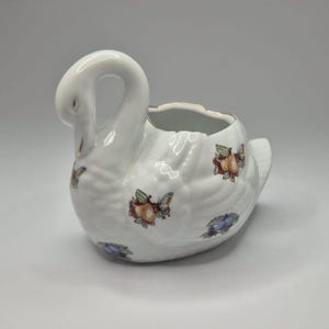 May include: White ceramic swan-shaped planter with a curved neck and open top. The planter is decorated with colourful butterfly and fruit motifs. The rim has a gold trim.