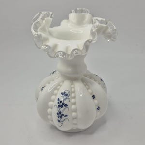 May include: A white glass vase with a frilled rim and a rounded body. The vase features blue floral patterns and a beaded texture. The frilled rim has a clear, translucent edge.