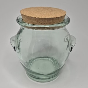 May include: A clear glass jar with a cork lid. The jar has two small handles on the sides and a rounded shape. The cork lid is circular and fits snugly on top of the jar.