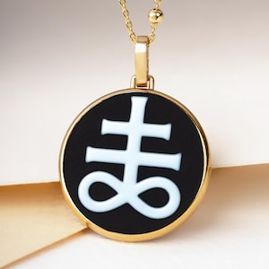 May include: Gold-toned necklace featuring a circular pendant with a black background and a white symbol. The pendant is framed in gold and hangs from a delicate chain with small gold beads.