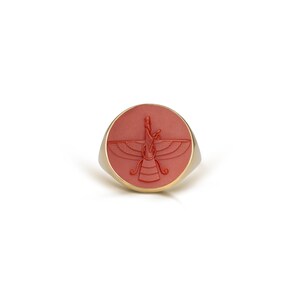 May include: A gold-toned ring with a round, coral-colored face. The face features a detailed, carved design of a winged figure. The ring has a simple, elegant design, suitable for everyday wear or special occasions.