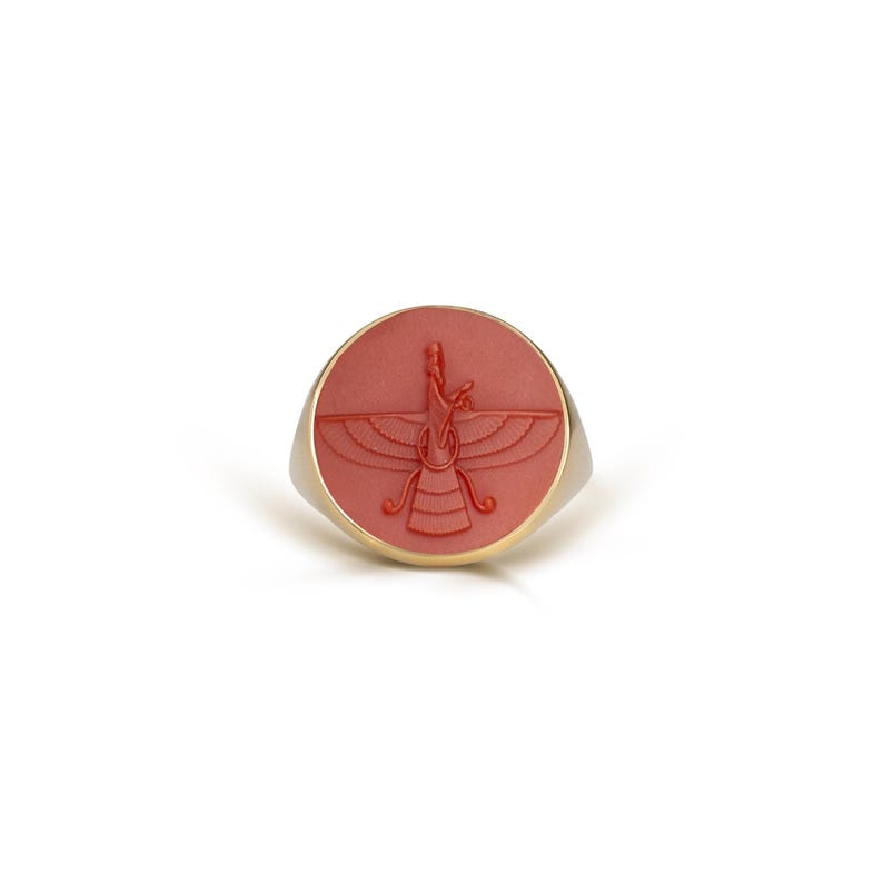 May include: A gold-toned ring with a round, coral-colored face. The face features a detailed, carved design of a winged figure. The ring has a simple, elegant design, suitable for everyday wear or special occasions.