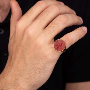 May include: A gold ring with a red, circular stone featuring an engraved design. The ring is worn on a finger, showcasing its polished gold band and the intricate details of the stone's artwork. The ring is a piece of jewelry.