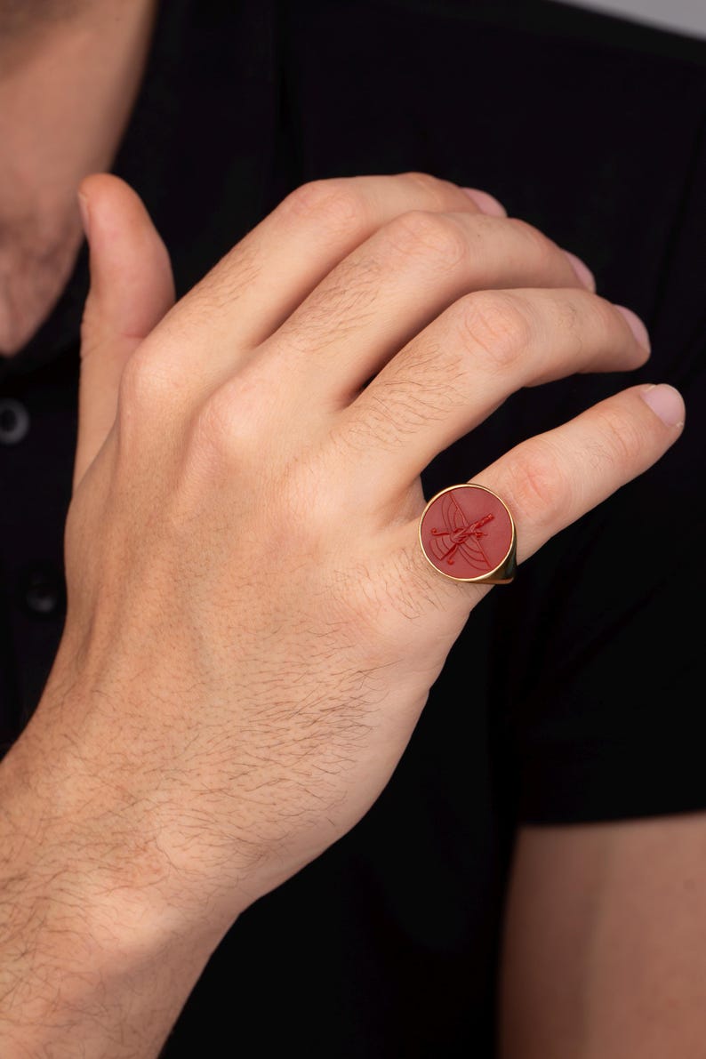 May include: A gold ring with a red, circular stone featuring an engraved design. The ring is worn on a finger, showcasing its polished gold band and the intricate details of the stone's artwork. The ring is a piece of jewelry.