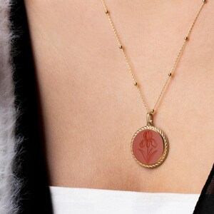 May include: A gold-colored necklace with a round pendant featuring a carved iris flower design in a reddish-brown hue. The pendant is encircled by a twisted rope design. The necklace is worn on a person.