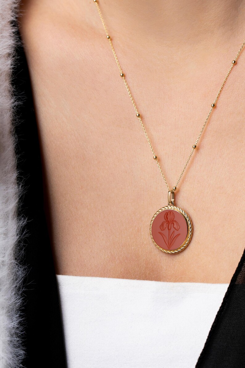 May include: A gold-colored necklace with a round pendant featuring a carved iris flower design in a reddish-brown hue. The pendant is encircled by a twisted rope design. The necklace is worn on a person.