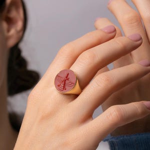 May include: A gold ring with a round, pinkish-red stone featuring a detailed engraved design. The ring is worn on a finger, showcasing the intricate craftsmanship and elegant style of the jewelry.