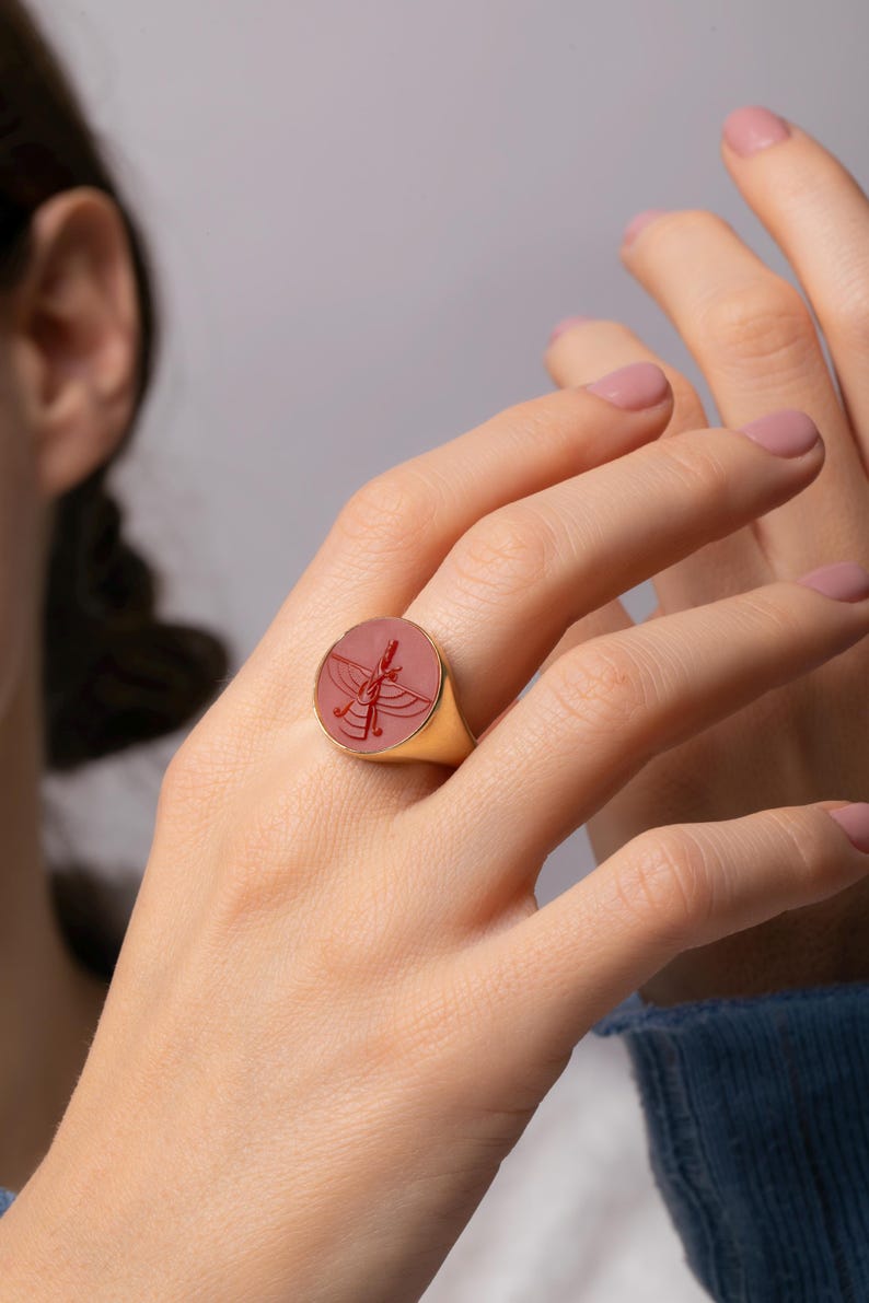 May include: A gold ring with a round, pinkish-red stone featuring a detailed engraved design. The ring is worn on a finger, showcasing the intricate craftsmanship and elegant style of the jewelry.