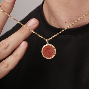 May include: Gold-toned necklace with a round pendant featuring a red iris design. The pendant is encircled by a twisted rope-like border. The necklace is held by a person wearing a black shirt.