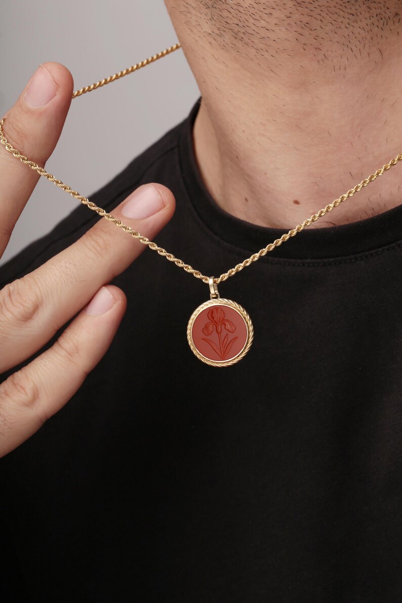 May include: Gold-toned necklace with a round pendant featuring a red iris design. The pendant is encircled by a twisted rope-like border. The necklace is held by a person wearing a black shirt.