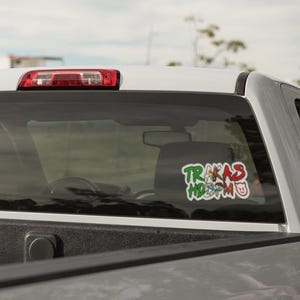 K&ouml;nnte beinhalten: Ein wei&szlig;er Pickup-Truck mit einem Aufkleber an der Heckscheibe, auf dem "TRAKAS HD3M0" steht. Der Aufkleber ist in Gr&uuml;n, Rot und Wei&szlig; mit einem Comic-Schwein.