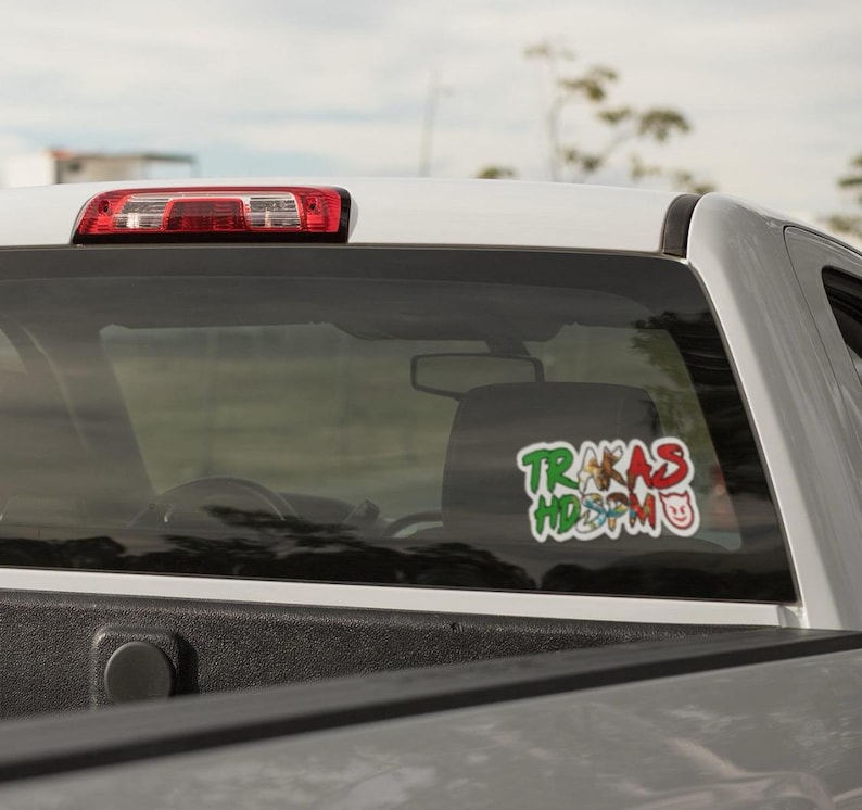 K&ouml;nnte beinhalten: Ein wei&szlig;er Pickup-Truck mit einem Aufkleber an der Heckscheibe, auf dem "TRAKAS HD3M0" steht. Der Aufkleber ist in Gr&uuml;n, Rot und Wei&szlig; mit einem Comic-Schwein.