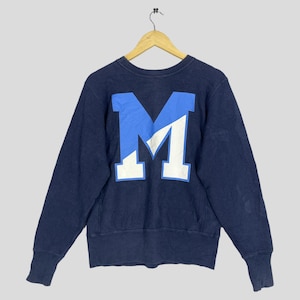 Op de afbeelding: Marineblauw sweatshirt met een grote, tweekleurige blauwe en witte letter "M" op de rug. Het sweatshirt hangt aan een houten hanger. Het heeft geribde manchetten en zoom.
