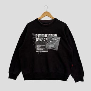 May include: Black sweatshirt with the text "PREDICTION REALZ" in white and gray. The sweatshirt features a graphic design with additional text and a signature. The garment is hanging on a wooden hanger.