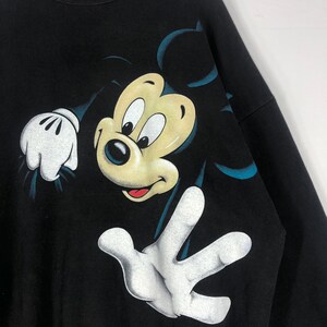 May include: Black sweatshirt featuring a large graphic of Mickey Mouse. The cartoon character is depicted with a yellow face, black eyes, and white gloves. The design includes blue accents and is set against a black background.