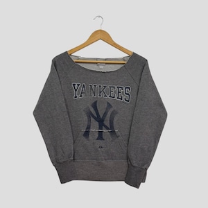 Può includere: Una felpa grigia con la scritta "YANKEES" in lettere blu navy sopra un logo "NY" blu navy. La felpa ha uno scollo a barchetta ampio e una tasca frontale. L'indumento è appeso a una gruccia di legno.