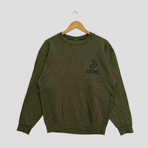 May include: Olive green sweatshirt with the text "USMC" and a graphic on the front. The long-sleeved crew neck top is hanging on a wooden hanger. The sweatshirt is made of a soft material.