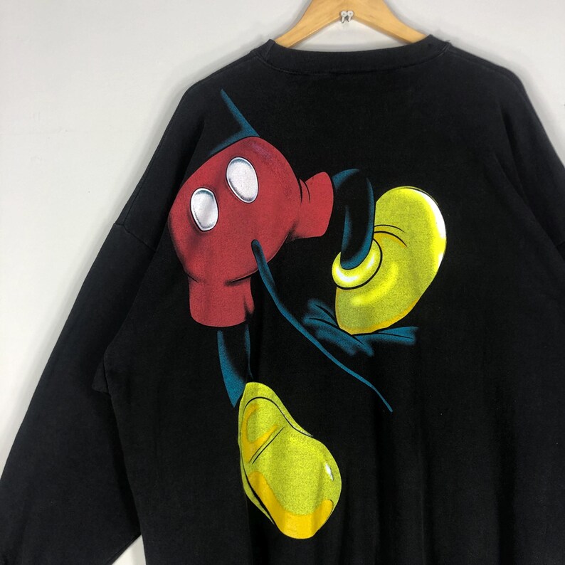 May include: Black long-sleeve sweatshirt featuring a large graphic of Mickey Mouse's red gloved hands and yellow shoes. The cartoon character's image is printed on the back of the shirt. The sweatshirt is hanging on a wooden hanger.