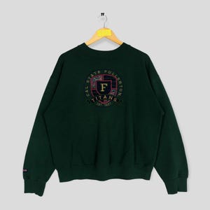 Könnte beinhalten: Grüner Pullover mit einem großen gestickten Logo auf der Rückseite. Das Logo zeigt einen Schild mit den Buchstaben "F" und "TITANS" sowie dem Text "CAL STATE FULLERTON EST. 1957".