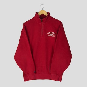 Puede incluir: Una sudadera roja con cuello alzado con cremallera y el texto "SAINT FRANCIS SFU UNIVERSITY" en blanco. La sudadera tiene mangas largas y dobladillos y puños acanalados. La prenda está colgada de una percha de madera.