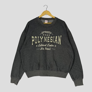 Może przedstawiać: Ciemnoszara bluza z napisem "AUTHENTIC POLYNESIAN Cultural Center Lāʻie, Hawaiʻi" w kremowym kolorze. Bluza ma okrągły dekolt i długie rękawy. Wisi na drewnianym wieszaku.