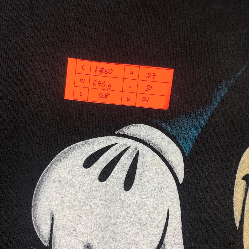 May include: Black garment featuring a white gloved hand and a portion of a character's face. An orange sticker with handwritten details is in the upper left corner. The garment's weight is 650 grams.