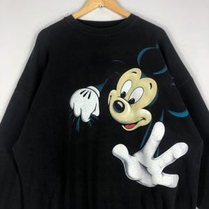 May include: Black sweatshirt featuring a large graphic of Mickey Mouse. The cartoon character's face is in the center, with his white gloved hands and arms extending to the sides. The sweatshirt is a classic crew neck style.