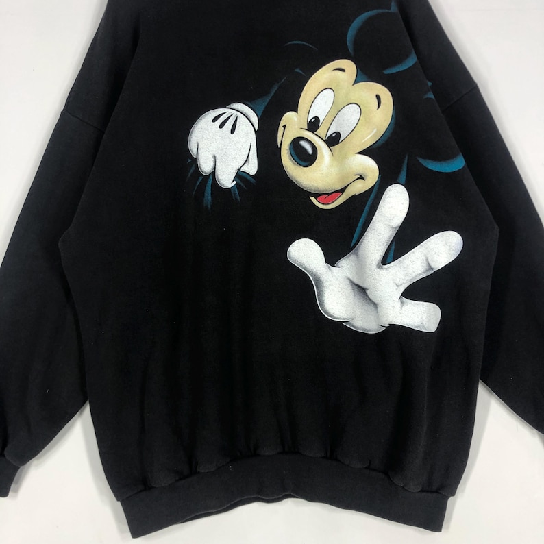 May include: Black sweatshirt featuring a large graphic of Mickey Mouse. The cartoon character's face and gloved hands are prominently displayed. The design includes white, yellow, red, and black colors. The sweatshirt has a crew neck and long sleeves.