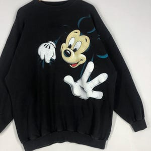 May include: Black sweatshirt featuring a large graphic of Mickey Mouse. The cartoon character is depicted with a cream-colored face, black eyes, and white gloves. The sweatshirt has a crew neck and long sleeves.