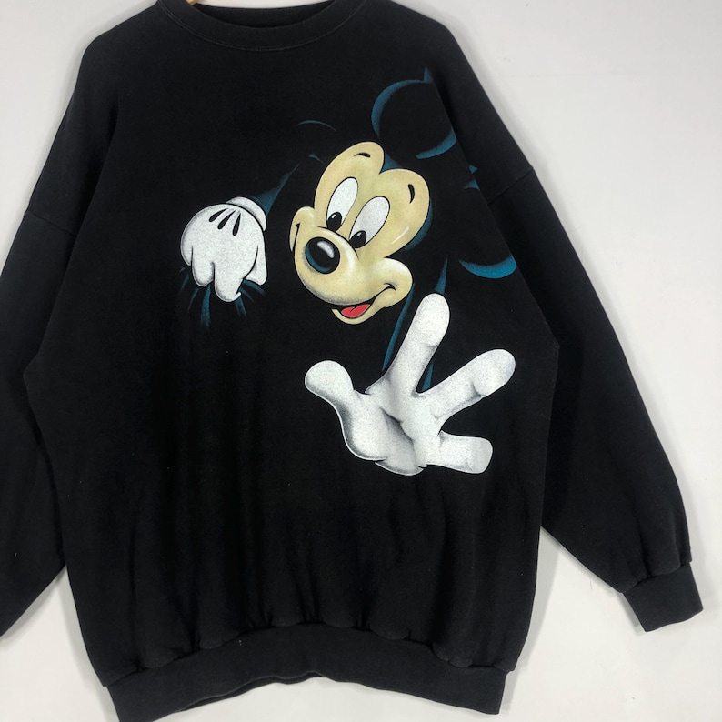 May include: Black sweatshirt featuring a large graphic of Mickey Mouse. The cartoon character is depicted with a cream-colored face, black eyes, and white gloves. The sweatshirt has a crew neck and long sleeves.