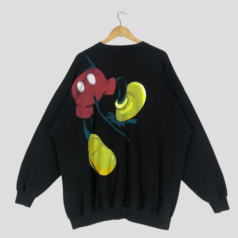 May include: Black sweatshirt featuring a large graphic of Mickey Mouse. The design shows Mickey's red gloved hands, yellow shoes, and black shorts. The sweatshirt is hanging on a wooden hanger.