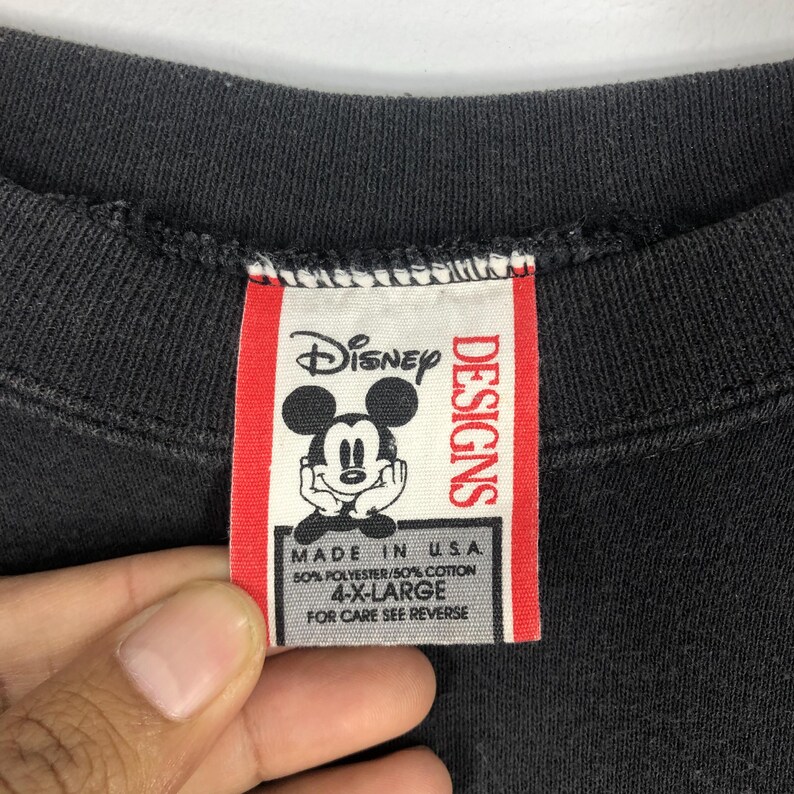 May include: Close-up of a black garment tag with the Disney logo featuring Mickey Mouse. The tag reads "DESIGNS" and "MADE IN U.S.A." with size "4-X-LARGE". The tag is red and white.