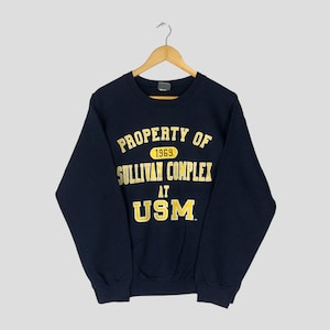 Puede incluir: Sudadera azul marino con el texto amarillo "PROPERTY OF SULLIVAN COMPLEX AT USM". El año "1969" está encima de "SULLIVAN COMPLEX". La sudadera está colgada de una percha de madera.