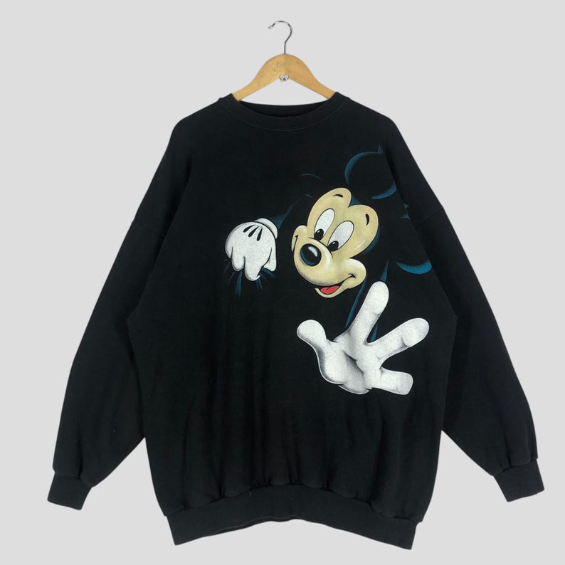 May include: Black sweatshirt featuring a large graphic of Mickey Mouse. The cartoon character is depicted with a white gloved hand reaching out. The sweatshirt has long sleeves and a crew neck.
