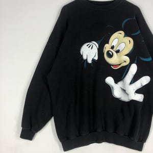 May include: Black sweatshirt featuring a large graphic of Mickey Mouse. The cartoon character's face and hands are prominently displayed in a playful pose. The design includes white gloves and a yellow face with a red mouth.