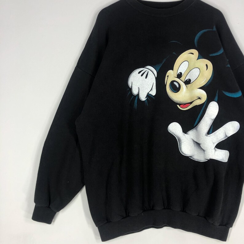 May include: Black sweatshirt featuring a large graphic of Mickey Mouse. The cartoon character's face and hands are prominently displayed in a playful pose. The design includes white gloves and a yellow face with a red mouth.