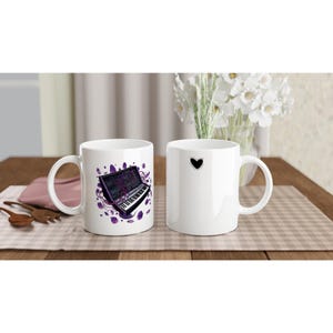 Può includere: Due tazze in ceramica bianca. Una tazza presenta un'illustrazione colorata di un sintetizzatore con accenti viola. L'altra tazza ha un design a cuore nero. Entrambe le tazze hanno un manico curvo e sono su una superficie a scacchi.