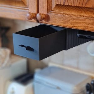 May include: A matte black under-cabinet storage drawer with a rectangular shape and a small horizontal handle on the front face. The drawer is mounted to the underside of a wooden cabinet and is shown in an open, extended position.
