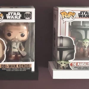 May include: Two Star Wars Pop! Vinyl figures. The figure on the left is Obi-Wan Kenobi, a light-skinned man with a brown beard and brown hair. The figure on the right is The Mandalorian with The Child, a green alien with large ears.