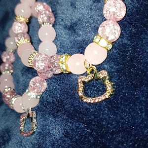 May include: Two pink and gold beaded bracelets with a gold charm in the shape of a cat. The charm has pink rhinestones.