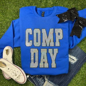 May include: Royal blue sweatshirt with the words "COMP DAY" in silver glitter lettering. The sweatshirt is laying on a pair of blue jeans with a black bow on the sleeve.